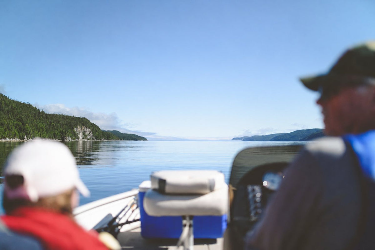Two friends in a fishing boat heading out across a calm lake lined with forested hills