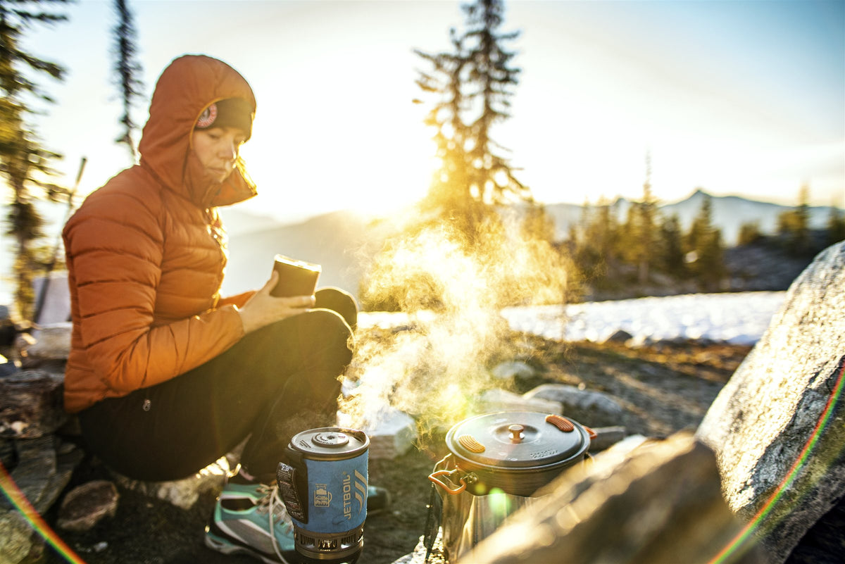 Hiker in an orange jacket making coffee on a camp stove at a mountain campsite
