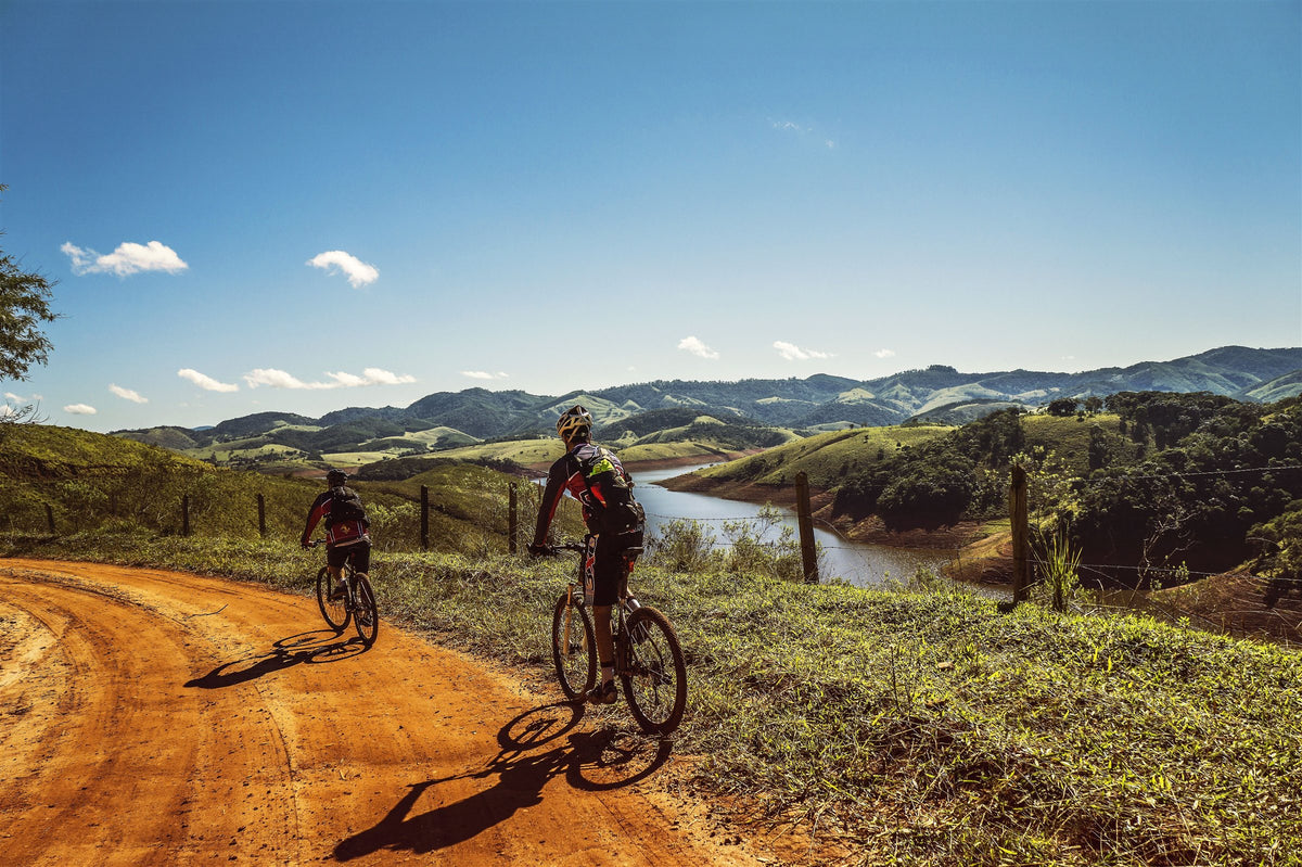 Two mountain bikers riding a red dirt road above a lake and green hills
